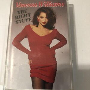 Puede incluir: Una cinta de casete con el álbum "The Right Stuff" de Vanessa Williams. La portada del álbum muestra a una mujer con un vestido rojo. El casete está dentro de una caja de plástico transparente.