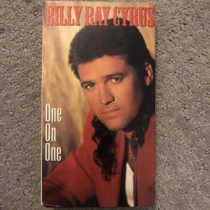 Puede incluir: Cinta de vídeo VHS del álbum "One On One" de Billy Ray Cyrus. La portada presenta un retrato de Cyrus con una chaqueta roja, con el título del álbum en blanco y el nombre del artista en rojo en la parte superior. El fondo es un degradado de colores cálidos.