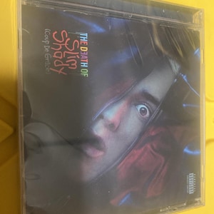 Puede incluir: Una caja de CD con el álbum "The Death of Slim Shady (Coup De Grâce)". La portada muestra un primer plano del rostro de una persona con una expresión intensa. El texto está en una fuente colorida y estilizada.
