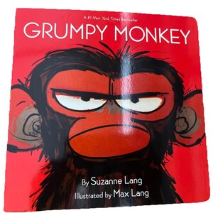 May include: A children's book cover with a red background and the title "GRUMPY MONKEY" in white. The illustration features a close-up of a grumpy-looking monkey with dark brown fur and a red face. The author and illustrator's names are also visible.