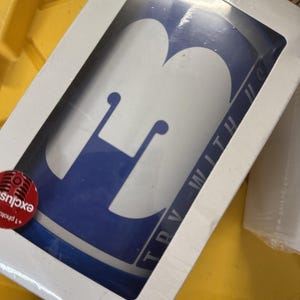 Puede incluir: Una taza azul y blanca con un gran diseño de "H" blanco. La taza está en una caja blanca con la inscripción "TRY WITH US" en el lateral. Una pegatina roja con la palabra "exclusive" es visible.