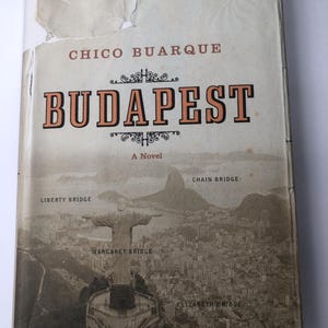 May include: A vintage copy of "Budapest: A Novel" by Chico Buarque. The book cover features the title in large orange letters with decorative accents. A black and white image of a cityscape with bridges and a statue is below the title.