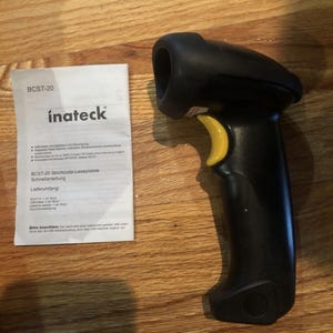 May include: A black barcode scanner with a yellow trigger and a black handle. The scanner is on a wooden surface. The text on the instruction manual reads "BCST-20 inateck".