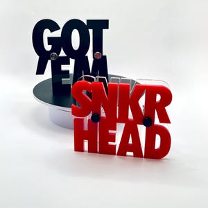 May include: Black and red acrylic signs with the phrases "GOT EM" and "SNKR HEAD". The black sign is on a black base, and the red sign is in front. The signs are on a white background.