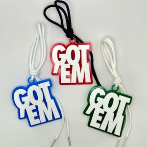 May include: Three decorative tags featuring the text GOT EM in bold white letters. One tag has a blue border, one has a red border, and one has a green border. Each tag is attached to a matching white or black cord loop for hanging or attachment.
