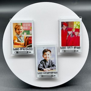 May include: A white circular display with three clear photo holders. Each holder contains a photo of a child and the text "hello preschool", "hello kindergarten", and "hello first grade" with a pencil graphic.