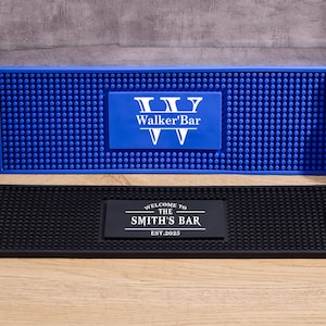 Könnte beinhalten: Zwei Barthekenmatten, eine blaue und eine schwarze, mit personalisiertem Text. Die blaue Matte hat "Walker'Bar" in Weiß. Auf der schwarzen Matte steht "WELCOME TO THE SMITH'S BAR EST.2025" in Weiß. Eine Bierflasche und eine Whiskyflasche sind sichtbar.