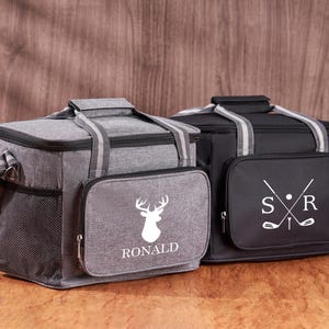 Puede incluir: Dos bolsas isot&eacute;rmicas personalizadas. Una bolsa es gris con una silueta de ciervo blanca y el nombre "RONALD" impreso en ella. La otra bolsa es negra con palos de golf blancos y las iniciales "S.R." impresas en ella.