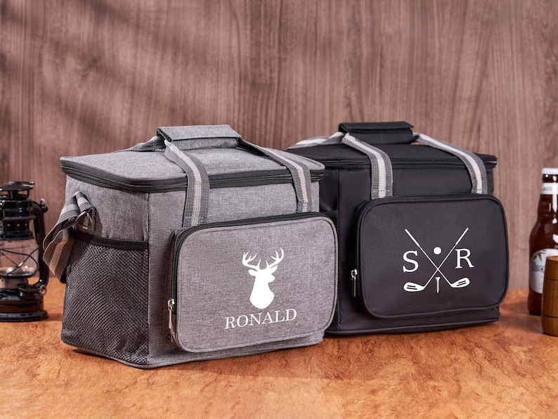 Puede incluir: Dos bolsas isot&eacute;rmicas personalizadas. Una bolsa es gris con una silueta de ciervo blanca y el nombre "RONALD" impreso en ella. La otra bolsa es negra con palos de golf blancos y las iniciales "S.R." impresas en ella.