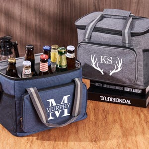 Puede incluir: Dos bolsas isot&eacute;rmicas personalizadas. La bolsa azul tiene un monograma blanco "M MURPHY" y la bolsa gris tiene un monograma blanco "KS" con cuernos de ciervo blancos. Ambas bolsas est&aacute;n llenas de botellas de cerveza.