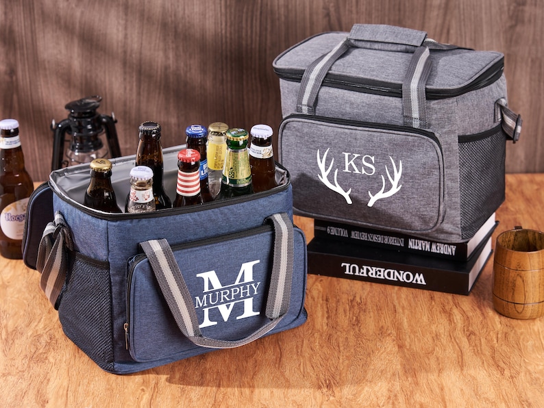 Puede incluir: Dos bolsas isot&eacute;rmicas personalizadas. La bolsa azul tiene un monograma blanco "M MURPHY" y la bolsa gris tiene un monograma blanco "KS" con cuernos de ciervo blancos. Ambas bolsas est&aacute;n llenas de botellas de cerveza.