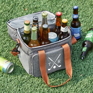 May include: A gray insulated cooler bag with brown handles and a white design of crossed golf clubs and the letters "T" and "R". The cooler bag is filled with various beer bottles and a can.