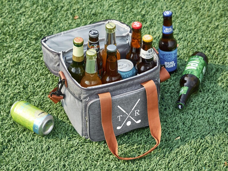 May include: A gray insulated cooler bag with brown handles and a white design of crossed golf clubs and the letters "T" and "R". The cooler bag is filled with various beer bottles and a can.
