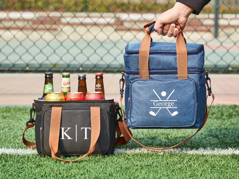 May include: Two personalized insulated cooler bags. One is black with brown straps and the initials "K|T" embroidered on the front. The other is blue with brown straps and the name "George" and a golf club design embroidered on the front.