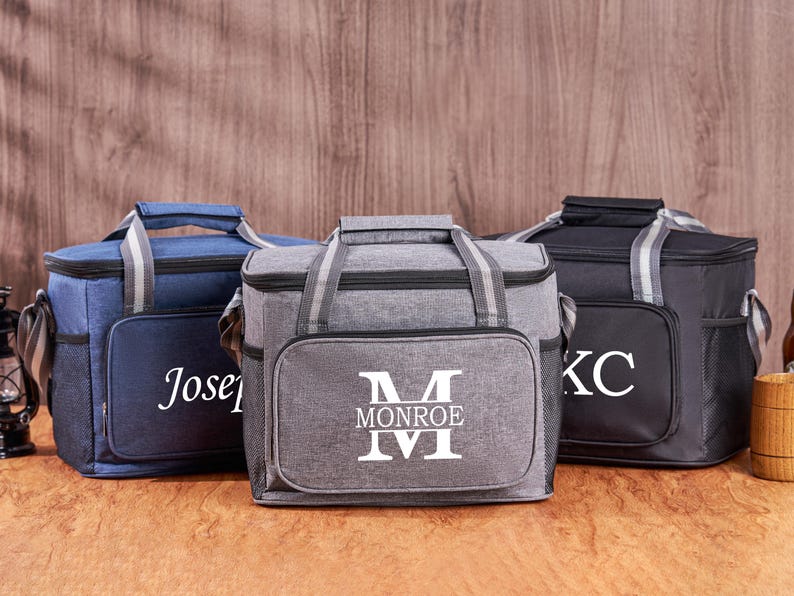 Puede incluir: Tres bolsas de almuerzo aisladas con nombres personalizados. Las bolsas son grises, azules y negras. La bolsa gris tiene un monograma blanco con el nombre "Monroe" y la inicial "M". La bolsa azul tiene el nombre "Joseph" bordado en blanco. La bolsa negra tiene las iniciales "KC" en blanco.