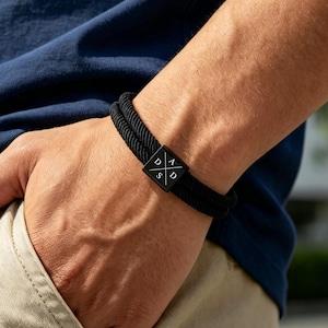 Op de afbeelding: Zwart touw armband met een vierkant, zwart metalen middelpunt. Het middelpunt heeft de letters D, A, S en D in het wit. De armband wordt om een pols gedragen, met een marineblauw overhemd en een beige broek op de achtergrond.
