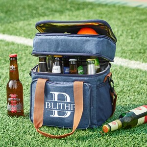 May include: A blue and brown personalized cooler bag with the letter "D" and the name "BLITHE" printed on the front. The cooler bag is filled with bottles of beer and has a brown handle.