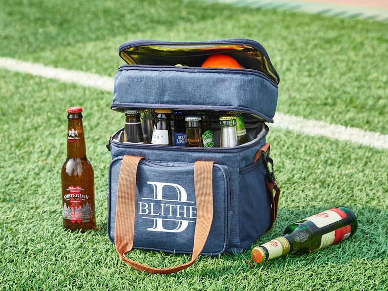 May include: A blue and brown personalized cooler bag with the letter "D" and the name "BLITHE" printed on the front. The cooler bag is filled with bottles of beer and has a brown handle.