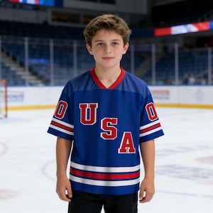 Pode incluir: Uma pessoa usando uma camisa de hóquei azul com as letras "USA" em vermelho. A camisa tem listras brancas e vermelhas nas mangas e na bainha. O fundo mostra uma pista de hóquei no gelo com um goleiro e espectadores.