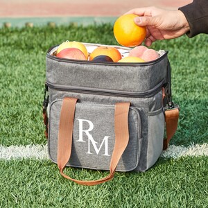 May include: A gray insulated lunch bag with brown handles and a white monogram "RM". The bag is filled with fruit, including oranges, apples, and plums.