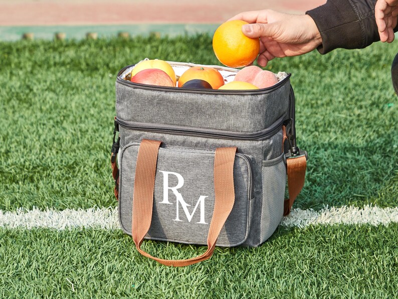 May include: A gray insulated lunch bag with brown handles and a white monogram "RM". The bag is filled with fruit, including oranges, apples, and plums.
