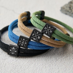 Puede incluir: Cuatro pulseras de cuerda en negro, azul, beige y verde. Cada pulsera tiene un dije cuadrado negro con símbolos y letras blancas. Las pulseras están apiladas sobre una superficie clara.