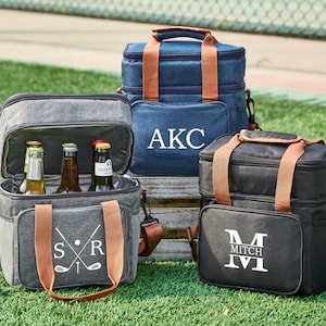 May include: Three personalized insulated cooler bags with brown leather handles. The bags are gray, blue, and black. The gray bag has a golf club design and the initials "SR". The blue bag has the initials "AKC". The black bag has the initial "M" and the name "Mitch".