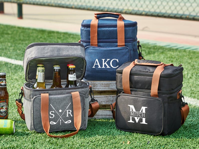 May include: Three personalized insulated cooler bags with brown leather handles. The bags are gray, blue, and black. The gray bag has a golf club design and the initials "SR". The blue bag has the initials "AKC". The black bag has the initial "M" and the name "Mitch".