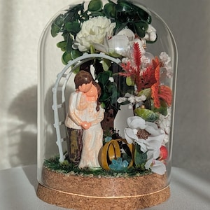 以下が含まれることがあります： コルク製の土台が付いたガラスのドームの中に、白いアーチと梯子のあるミニチュアのシーンがあります。アーチの下にはカップルが立っており、緑と赤の花に囲まれています。手前に2つの金の指輪が見えます。