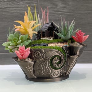 May include: A miniature black house with a thatched roof sits on a bed of green moss in a black, ornate planter. The planter is decorated with a swirling pattern and has a raised lip. The house is surrounded by a variety of succulents and small plants, including a pink succulent and a yellow succulent.