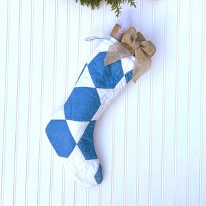 May include: A quilted Christmas stocking with a blue and white geometric pattern. The stocking is decorated with a burlap bow and hangs against a white paneled wall. A small sprig of greenery is at the top.