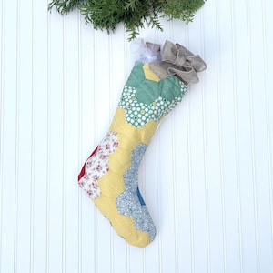 May include: A handmade Christmas stocking with a patchwork design. The stocking features a variety of fabrics in yellow, green, red, and blue, with a gray bow at the top. The stocking is decorated with floral and polka dot patterns.