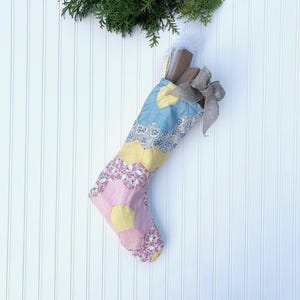May include: A handmade Christmas stocking with a patchwork design in pastel colors. The stocking is filled with gifts and tied with a gray ribbon. The background is a white wall with vertical lines, and a sprig of greenery is at the top.