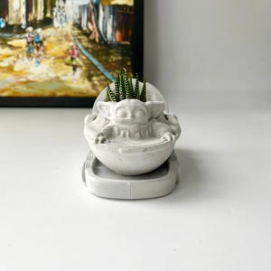 May include: A white ceramic planter shaped like a character from the Star Wars franchise, holding a small green succulent plant. The planter is sitting on a white ceramic base.