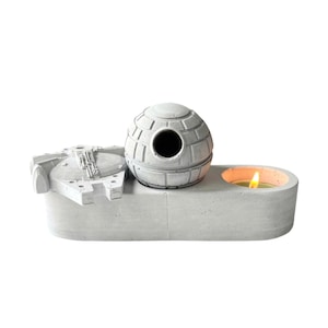 May include: A gray concrete candle holder shaped like a spaceship with a Death Star on top. The candle holder has a small, round candle holder on the side.