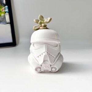 May include: A white 3D-printed Stormtrooper helmet planter with a small succulent plant. The planter is a detailed replica of the iconic Star Wars character's helmet, perfect for small plants. The succulent has green and tan leaves.