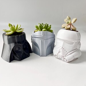 May include: Three 3D-printed planters shaped like Star Wars characters: Darth Vader in black, a Mandalorian helmet in gray, and a Stormtrooper helmet in white. Each planter holds a small succulent plant, adding a touch of greenery.