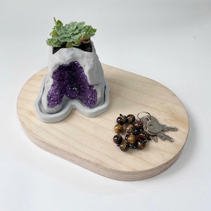 May include: A small succulent plant growing in a concrete planter shaped like a purple amethyst geode. The planter is sitting on a wooden tray with a set of keys and a beaded necklace.