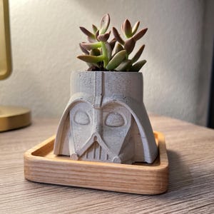 May include: A white ceramic planter shaped like Darth Vader's head, with a succulent plant growing inside. The planter is sitting on a wooden tray.