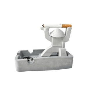 May include: A gray concrete ashtray with a unique design. A small figure holds a lit cigarette. The ashtray has a rectangular shape with a recessed area for ashes and a curved indentation for resting cigarettes. The design is modern and minimalist.