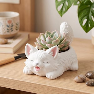 May include: A white ceramic planter shaped like a smiling cat with textured fur, pink-tinted inner ears, and an open mouth. The cat is in a lying position with its tail raised, holding a green and pink succulent plant in its back section.