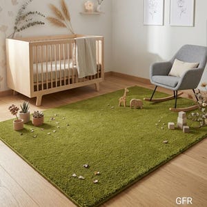 May include: A rectangular, solid olive green area rug with a plush, textured surface. The rug is positioned on a light-coloured wooden floor in a room containing a wooden crib, a grey rocking chair, small wooden animal figures, and blocks.