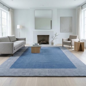 May include: A large rectangular area rug featuring a solid deep blue centre panel framed by a wide light grey border. The rug has a low-pile texture and is positioned centrally in a room, covering a significant portion of the light-coloured wooden floor.