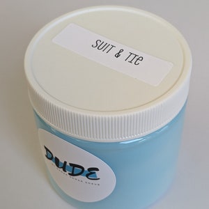 May include: A light blue jar of sugar scrub with a white lid and a label that reads "SUIT & TIE." The jar has a white label with the word "DUDE" in a stylized font. The product is likely a skincare item.