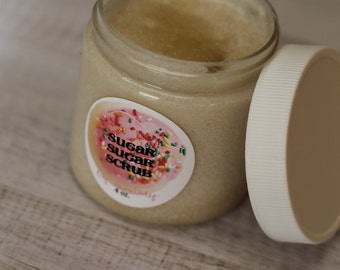 May include: An open glass jar of sugar scrub with a white lid. The jar has a label with the words "Sugar Sugar Scrub" and a pink and colorful design. The jar contains a light brown scrub and is 4 oz.