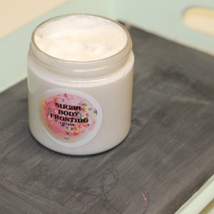 May include: A jar of Sugar Body Frosting Lotion sits on a gray surface. The jar is white with a round label featuring a pink donut design and the product name. The lotion inside is white and fluffy.