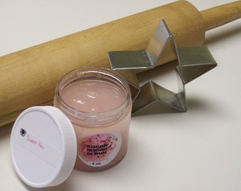 May include: A clear glass jar with a pink sugar scrub inside. The jar has a white lid with the words "Sweet Pea" written on it. The jar is sitting on a white surface next to a wooden rolling pin and a metal star-shaped cookie cutter.