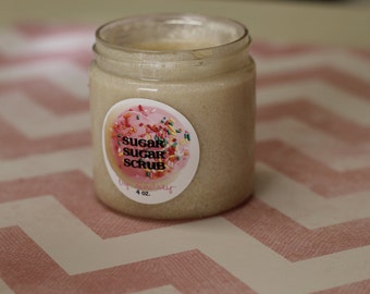 May include: A clear glass jar filled with a beige sugar scrub. The jar has a circular label with the words "Sugar Sugar Scrub" in pink and yellow, decorated with sprinkles. The label also says "4 oz."