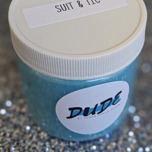 May include: A clear jar with a blue glitter slime inside. The jar has a white lid with a label that says "SUIT & TIE". The jar is sitting on a sparkly silver surface. The label on the jar also says "DUDE BY SUGAR SUGAR CREW".