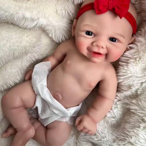 May include: A realistic baby doll with fair skin, blue eyes, and a red bow headband. The doll is wearing a white nappy and is lying on a fluffy, white blanket. The doll has a smiling expression.
